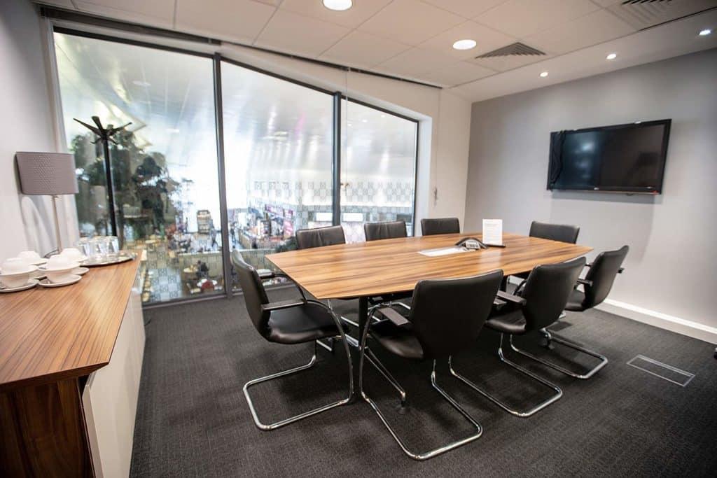 Modern conference room with a large wooden table, black chairs, and a television on the wall, featuring a city view through the window and Drik catered food on the table.