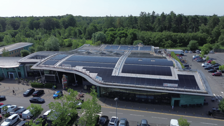Aerial view of a shopping complex with a curved roof covered in solar panels, with a parking lot and dense green trees behind. “Pizza Express” storefront visible at the front.