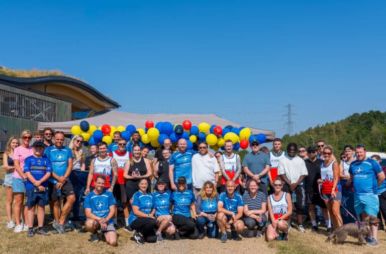 Group photo of volunteers in blue uniforms posing outdoors under a clear blue sky with balloons and a tent behind them.