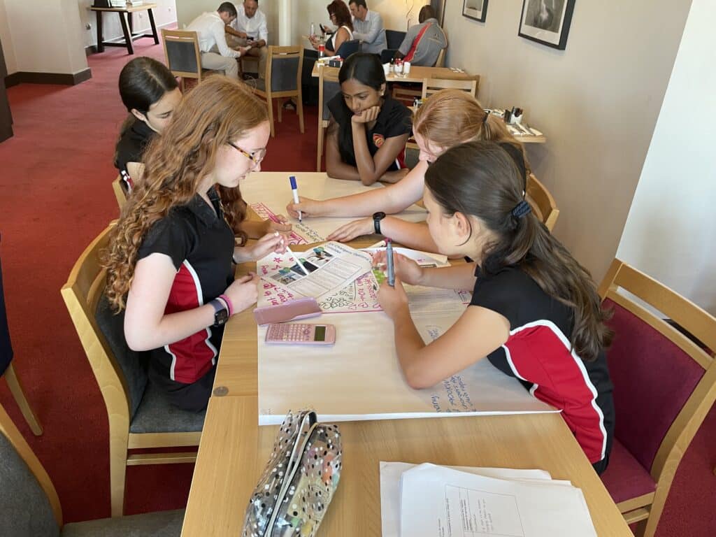 Group of students collaboratively writing on a large poster at a wooden table in a cafe-like study area.