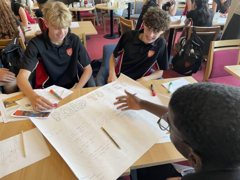 Students in black polo shirts collaborate at a table, drawing and writing on a large poster with markers in a bright classroom.