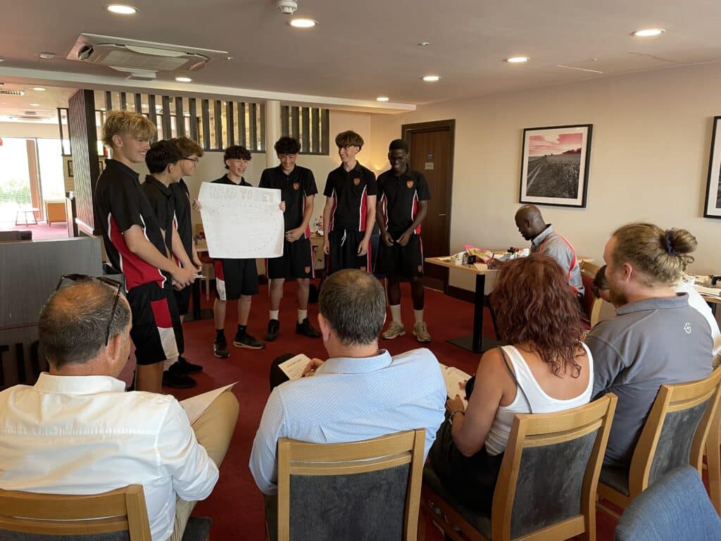 Six male students in matching black and red uniforms present a large poster to an audience in a meeting room.